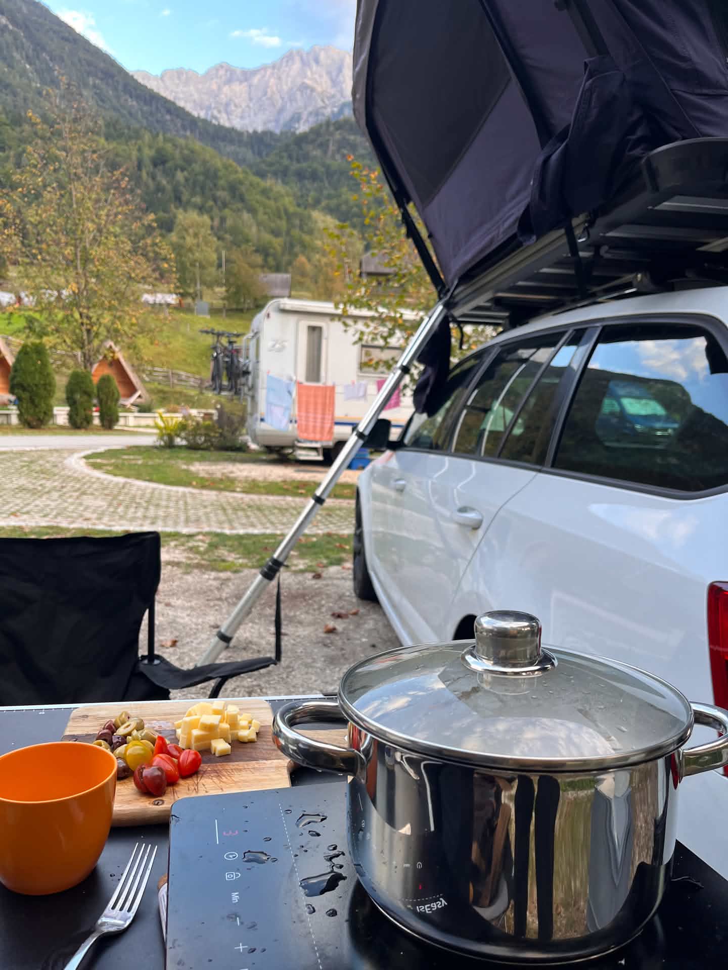 Kochen am Auto mit aufgeklapptem Dachzelt auf dem Campingplatz, im Hintergrund Berge